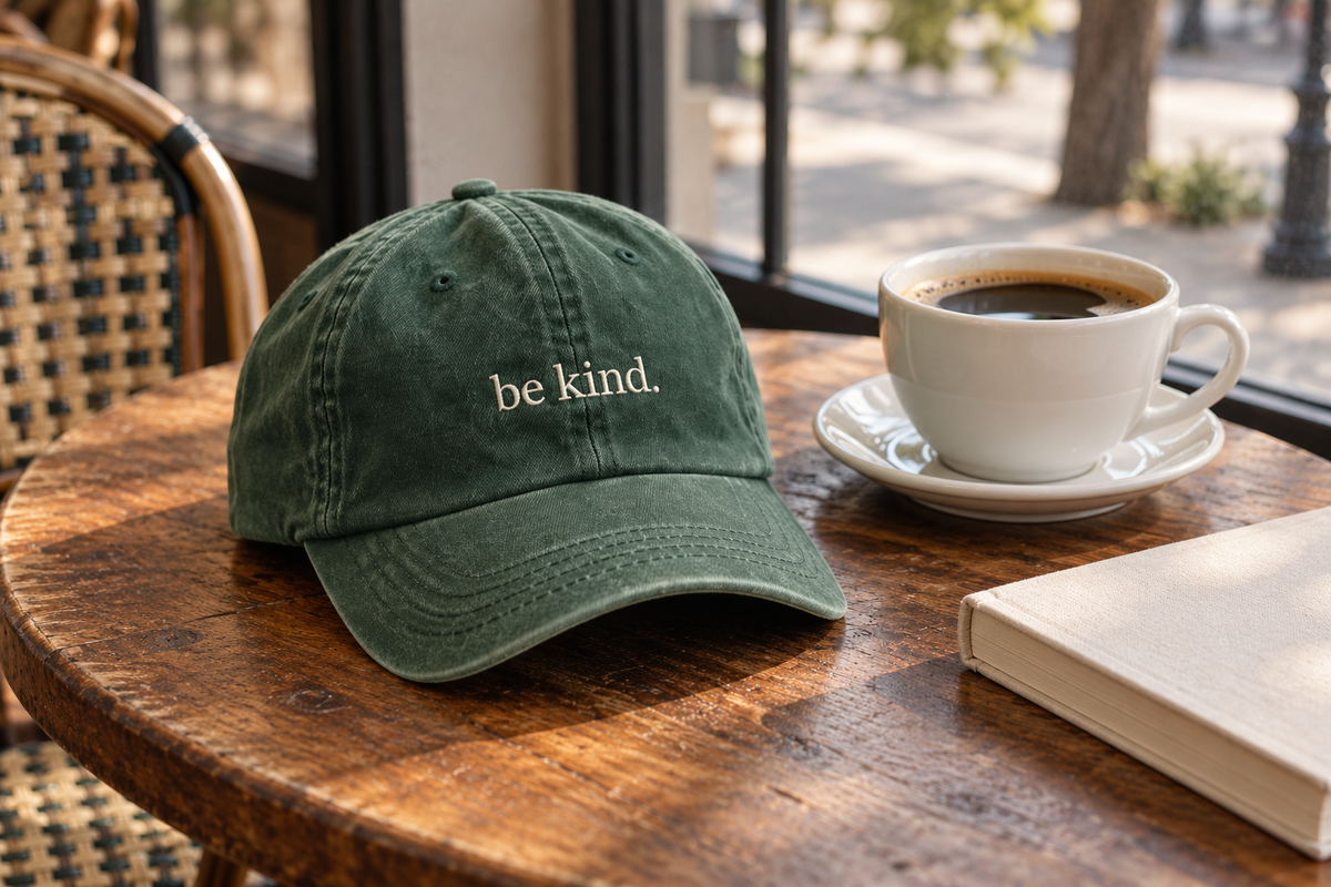 AI-generated Fika editorial: pine Be Kind cap beside coffee and a book on a cafe table.