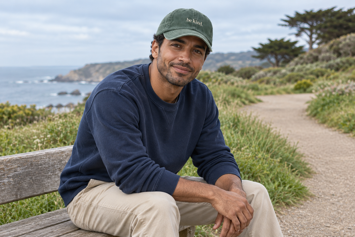 AI-generated Fika editorial: man wearing a pine Be Kind cap beside a coastal path.
