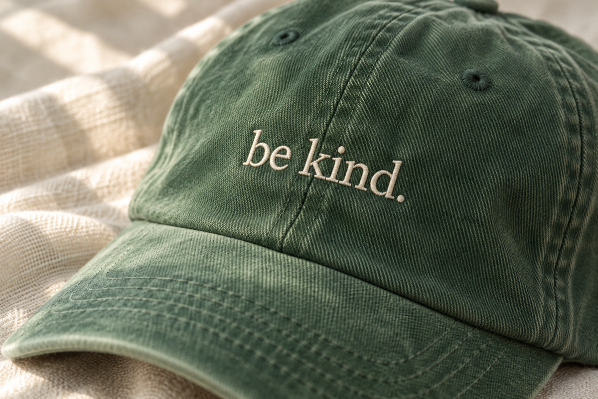 AI-generated Fika editorial close-up of ivory be kind embroidery on a pine cap.